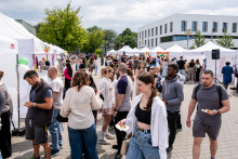 Tłum ludzi na plenerowym wydarzeniu. W tle rzędy białych namiotów wystawowych i nowoczesny budynek z dużymi oknami. Uczestnicy ubrani różnorodnie, niektórzy trzymają talerze z jedzeniem. Wydarzenie odbywa się na wybrukowanym placu otoczonym zielenią. Pogoda pochmurna, ale jasna.