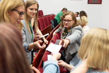 Grupa osób w sali konferencyjnej z czerwonymi krzesłami. Część osób stoi, część siedzi, przekazując sobie kartki papieru. W tle widoczne są inne osoby oraz fragmenty ścian z ramkami.