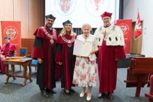 Cztery osoby, w tym trzy w togach akademickich i biretach. W centrum kobieta w białej sukience w kwiaty trzyma dyplom. W tle dwa banery i ekran z logo uczelni. Na pierwszym planie po lewej stronie stół i krzesło.