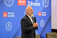 Mężczyzna w garniturze przemawia, trzymając mikrofon, na tle niebieskiej ścianki z logotypami „Instytut Ochrony Zdrowia” i „Szczyt Zdrowie”. Na ściance widoczne dwa czerwone prostokąty z białymi strzałkami skierowanymi w górę.