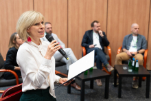 Kobieta z mikrofonem i kartką stoi przed czterema osobami siedzącymi w półokręgu. Przed nimi stoliki z butelkami wody. W tle drewniana ściana. 