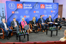 Siedem osób siedzi na scenie podczas panelu dyskusyjnego. Tło stanowi niebieska ścianka z logotypami „Szczyt Zdrowie” i symbolami medycznymi. Uczestnicy ubrani formalnie, przed nimi dwa stoliki z butelkami wody. Jeden z mężczyzn trzyma mikrofon i prowadzi rozmowę.