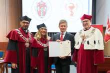 Cztery osoby stoją obok siebie, trzy w bordowych togach akademickich z łańcuchami. Mężczyzna pośrodku trzyma dyplom, jest ubrany w garnitur i czerwony krawat. W tle ekran z logo uczelni medycznej i flaga z herbem.