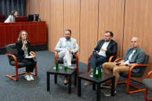 Trzech mężczyzn siedzi w półokręgu na panelu dyskusyjnym. Przed nimi dwa stoliki z butelkami wody. Po lewej kobieta z mikrofonem prowadząca panel. W tle drewniana ściana, biurko z komputerem i osoba siedząca za monitorem.