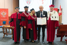 Pięć osób pozuje do zdjęcia w auli. Cztery w czerwono-czarnych togach i biretach, jedna w garniturze trzyma dyplom. W tle dwa banery i ekran z logo uczelni, po prawej czerwona flaga z herbem.