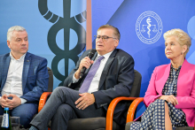Trzy osoby siedzą na krzesłach. Mężczyzna po lewej ma siwe włosy, niebieską marynarkę i białą koszulę. Mężczyzna w środku trzyma mikrofon, ubrany jest w ciemny garnitur i fioletowy krawat. Kobieta po prawej ma różową marynarkę i sukienkę w czarno-biały wzór. W tle logotypy związane z ochroną zdrowia i symbol kaduceusza.