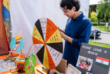 Młody mężczyzna stoi przy stoisku z rękodziełem. Ma ciemne włosy i niebieską koszulę. Na pierwszym planie kolorowa ruletka z angielskimi napisami, m.in. ‘Pick with your eyes closed’. Obok ozdobne pudełka, obrazki i tablica z flagą Indii. 