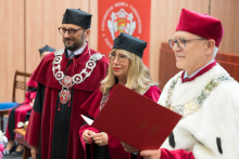 Trzy osoby w tradycyjnych togach akademickich. Dwie w czerwonych togach z łańcuchami, jedna w białej todze z czerwonym biretem. Jedna osoba trzyma czerwoną teczkę. W tle baner z herbem wydziału i fragment sali.