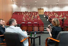 Aula z widownią na czerwonych fotelach. Na pierwszym planie trzech prelegentów siedzi i prowadzi dyskusję, jeden trzyma mikrofon. Przed nimi stolik z butelkami wody. W tle widoczne schody i wyjście ewakuacyjne.