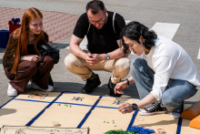 Trzy osoby siedzą na macie rozłożonej na chodniku i biorą udział w grze planszowej. Mata podzielona jest na kwadraty i zawiera pionki oraz karty.