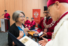 Wręczenie odznaczeń. Kobieta odbiera medal od mężczyzny w stroju akademickim z łańcuchem rektorskim. Obok drugi mężczyzna w todze. W tle osoby w czerwonych togach i biretach oraz mównica z logo uczelni. Na ścianie baner z herbem i napisem „Warszawski Uniwersytet Medyczny"..