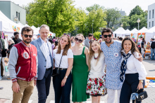 Siedem osób stoi na zewnątrz podczas wydarzenia. Są to władze dziekańskie i przedstawiciele samorządów studenckich.  tle namioty wystawowe i drzewa. Osoby ubrane różnorodnie: m.in. mężczyzna w czerwonej kurtce, kobieta w białej bluzce i zielonej sukience, kobieta w kwiecistej spódnicy oraz kobieta z apaszką na szyi.