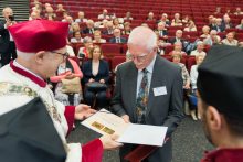 Uroczystość wręczenia dyplomów w sali z czerwonymi fotelami. Osoba w stroju ceremonialnym z biretem i peleryną wręcza dyplom starszemu mężczyźnie w garniturze. W tle widoczna publiczność, głównie osoby starsze.