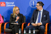 Dwie osoby siedzą na krzesłach podczas konferencji. Kobieta po lewej trzyma mikrofon i notatnik, ubrana w czarny garnitur. Mężczyzna po prawej w granatowym garniturze, białej koszuli i niebieskim krawacie. W tle niebieska ścianka z logotypem Instytutu Ochrony Zdrowia i napisem „Szczyt Zdrowie”.