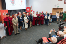 Grupa osób pozuje do zdjęcia w sali konferencyjnej. W centrum osoby w togach i biretach. W tle dwa ekrany z logo uczelni i flaga Polski. Po prawej dwie osoby siedzą i robią zdjęcia, po lewej mównica z herbem WUM.