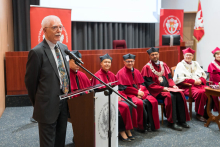 Mężczyzna w garniturze przemawia przy mównicy z mikrofonem. W tle sześć osób w czerwonych togach i biretach, dwie w białych togach z łańcuchami rektorskimi. Widoczne czerwone banery z logo uczelni.