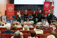 Występ Chóru WUM. Pięć osób stoi na scenie w audytorium, w tym kobieta trzymająca dziecko. Wszyscy ubrani elegancko. Przed sceną publiczność robi zdjęcia telefonami. W tle dwa czerwone banery z logo Warszawskiego Uniwersytetu Medycznego i napisami „Wydział Lekarski” oraz „Warszawski Uniwersytet Medyczny”.