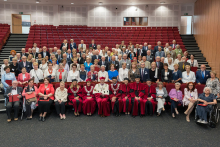 Około 100 osób pozuje do wspólnego zdjęcia w audytorium. W pierwszym rzędzie siedzą osoby w czerwonych togach akademickich i osoby starsze. Za nimi stoją pozostali uczestnicy na tle czerwonych krzeseł. W tle widoczne są schody i drzwi.