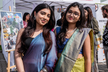 Dwie młode kobiety w tradycyjnych strojach stoją obok siebie. Jedna ma niebiesko-fioletowe sari, druga zielono-żółte sari z niebieską bluzką. Obie mają długie ciemne rozpuszczone włosy. W tle stoiska z plakatami i banerami, w tym z napisami ‘Pakistan’ oraz ‘Groceries and more from Pakistan'.