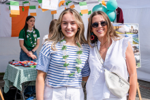 Dwie młode kobiety z zielonymi girlandami na szyjach stoją przed stoiskiem z irlandzkimi flagami, balonami i plakatem z krajobrazami. Jedna osoba w biało-niebieskiej bluzce w paski i białych spodniach, druga w białej bluzce bez rękawów z jasną torebką. Obie mają jasne włosy. Uśmiechają się do zdjęcia.