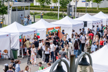 Rząd białych namiotów, wokół których gromadzą się ludzie. Uczestnicy rozmawiają i biorą udział w różnych aktywnościach. Niektóre namioty ozdobione balonami i banerami. W tle drzewa i budynki.