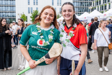 Dwie młode kobiety stoją obok siebie, trzymając kije do hurlingu. Jedna ma zieloną koszulkę Vodafone i białe spodenki, druga czerwoną koszulkę z białymi elementami i niebieskie spodenki. Obie mają na szyjach kwiatowe girlandy. W tle ludzie i namioty.