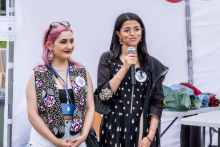 Dwie młode kobiety stoją obok siebie na tle białego namiotu. Jedna ma różowe włosy, kolorową kamizelkę z cekinami i biały top, druga czarną sukienkę z białym wzorem i trzyma mikrofon, do którego przemawia. W tle widać biały namiot.