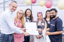 Sześć osób stoi pod namiotem na zewnątrz. Trzy osoby po lewej trzymają wspólnie dyplom, który wręczają osobie po prawej. Twarze zostały zamazane. W tle balony w kolorach białym, różowym i żółtym.