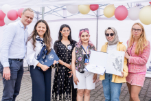 Sześć osób stoi pod białym namiotem na brukowanej powierzchni. W tle balony w kolorach czerwonym, białym i żółtym. Jedna osoba trzyma otwartą teczkę z dokumentami i dyplomem. Stroje osób są różnorodne, od eleganckich po casualowe. 