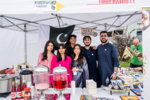 Grupa osób - młodych kobiet i młodych mężczyzn stoi pod białym namiotem z flagą Pakistanu. Uśmiechają się do zdjęcia. Na stole przed nimi znajdują się dwa dyspensery z różowymi napojami, butelki z sosami, książki i inne przedmioty. 