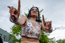 Młoda kobieta z ciemnymi rozpuszczonymi włosami topie z kwiatowym wzorem i przezroczystym panelem na dekolcie unosi ręce, wskazując palcami do góry. Na nadgarstkach bransoletki, w uszach kolczyki. W tle drzewa i fragment budynku.