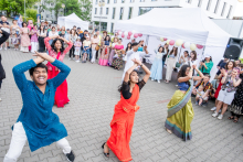 Młode kobiety i młodzi mężczyźni w tradycyjnych strojach tańczą na wybrukowanym placu przed nowoczesnym budynkiem. Twarze są zasłonięte. W tle tłum ludzi, część fotografuje lub nagrywa. Widoczne są białe namioty i balony w kolorach różowym, białym i złotym.