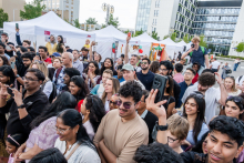 Tłum ludzi zgromadzonych na zewnątrz przed białymi namiotami wystawowymi. W tle nowoczesne budynki. Część osób robi zdjęcia lub nagrywa filmy telefonami. 