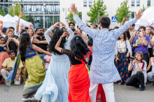 Grupa osób w kolorowych, tradycyjnych strojach tańczy na brukowanej powierzchni podczas wydarzenia plenerowego. W tle tłum obserwatorów robiących zdjęcia, namioty i budynki biurowe. Scena odbywa się na zewnątrz.