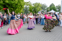 Trzy kobiety tańczą na świeżym powietrzu w kolorowych, tradycyjnych strojach. Jedna ma różową spódnicę i czerwony wachlarz, druga fioletową spódnicę z czerwoną szarfą, trzecia czarną spódnicę i różową bluzkę z niebieskim wachlarzem. W tle widzowie, drzewa i namiot.