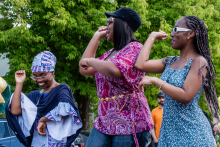 Trzy kobiety tańczą na świeżym powietrzu. Jedna ma tradycyjny afrykański strój i chustę na głowie, druga różową bluzkę ze wzorem i czarną czapkę, trzecia niebieską sukienkę w białe kropki. W tle drzewa, samochody i inne osoby.