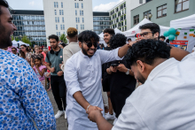 Dwóch mężczyzn w tradycyjnych strojach tańczy, trzymając się za ręce, podczas wydarzenia. W tle widoczne są inne osoby, namioty i budynki mieszkalne. Część uczestników rozmawia lub korzysta z telefonów. Na niebie chmury