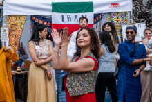 Grupa osób stoi przed stoiskiem z napisem „IRAN” na tle flagi Iranu. Na pierwszym planie kobieta w czerwonej bluzce i kamizelce klaszcze. Część osób ubrana w tradycyjne stroje. W tle kolorowe dekoracje i uczestnicy wydarzenia.