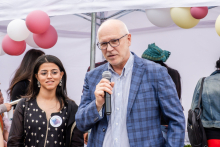 Mężczyzna - rektor WUM - w niebieskiej marynarce w kratę trzyma mikrofon i przemawia. Obok stoi młoda kobieta z długimi ciemnymi włosami w czarnej sukience z białymi guzikami. W tle balony w kolorach białym, różowym i żółtym oraz uczestnicy wydarzenia.