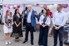 Pięć osób stoi pod białym namiotem na zewnątrz. Mężczyzna w środku - rektor WUM - trzyma mikrofon i przemawia. W tle balony w kolorach białym, różowym i żółtym oraz inni uczestnicy wydarzenia. Podłoże brukowane.