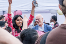 Grupa ludzi na zewnątrz podczas wydarzenia. Kobieta w czerwonej sukience z wzorami unosi telefon, robiąc zdjęcie lub film. Na jej rękach widoczna henna. Obok stoi mężczyzna w niebieskiej marynarce. W tle kolorowe balony i uczestnicy wydarzenia.