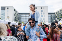 Tłum ludzi na zewnątrz podczas festiwalu. Młody mężczyzna w koszuli trzyma na ramionach dziewczynkę w niebieskiej bluzie i różowych spodniach. W tle nowoczesne budynki i białe namioty wystawowe, wokół osoby oglądające stoiska i rozmawiające.