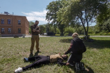 On the grass, three people participate in the simulation. One is lying in a tactical vest, another is kneeling next to him and performing rescue operations, and the third is standing with his hand at his chin. In the background is a brick building, trees and bushes.