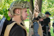A group of people wearing military helmets and protective vests stand in the open air surrounded by trees. Faces are blurred for anonymity. Green trees are visible in the background.