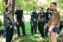 Six people in military clothing stand on the grass in the park. Three of them are holding replica weapons. Trees and shrubs are visible in the background. 
