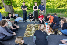 On the grass, surrounded by trees, a paramedic in an orange and black outfit is giving aid to a person wrapped in a gold rescue wrap. About a dozen people in dark clothing are around, some sitting, some standing. Green vegetation is visible in the background.