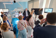 Grupa osób uczestniczy w prezentacji w sali Centrum Symulacji Medycznych. W centrum łóżko szpitalne z niebieskim pokryciem, otoczone sprzętem medycznym. Na ścianach dwa ekrany z obrazami medycznymi. Uczestnicy stoją wokół łóżka, skupieni na osobie prowadzącej przy ekranie.