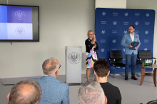 Na pierwszym planie osoby siedzące tyłem. Na środku sali kobieta, prorektorka, z mikrofonem i mężczyzna z dokumentami stoją obok mównicy z logo. Kobieta jest ubrana w kolorową sukienkę i czarny sweter. Mężczyzna ma na sobie niebieską marynarkę, białą koszulę i ciemne spodnie. W tle ekran i niebieska ścianka z powtarzającym się logo i tekstem.