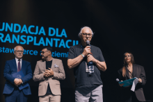 Na scenie stoją cztery osoby: trzech mężczyzn i jedna kobieta. Mężczyzna po lewej ma niebieski garnitur i trzyma statuetkę. Obok niego mężczyzna w beżowym garniturze. Dalej starszy mężczyzna w czarnej koszulce z mikrofonem. Kobieta po prawej w ciemnym stroju trzyma mikrofon i teczkę. W tle napis „FUNDACJA DLA TRANSPLANTACJI”.