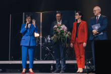 Na scenie stoją cztery osoby. Kobieta po lewej ma niebieski garnitur i czerwone buty, trzyma mikrofon. Obok niej mężczyzna w ciemnym garniturze z bukietem kwiatów. Dalej kobieta w czerwonym garniturze. Po prawej mężczyzna w ciemnym garniturze z teczką z logo uczelni. W tle perkusja i instrumenty za przezroczystymi osłonami.